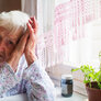 Bildet viser en trist, gammel dame som lener hodet mot hendene sine. Hun sitter ved kjøkkenbordet med et pilleglass foran seg.