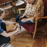 Bildet viser en hjemmesykepleier som skifter strømper på en eldre dame som sitter i stolen i hjemmet sitt.