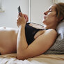 Bildet viser en gravid kvinne som ligger i BHen på senga. Hun kikker på mobilen og ser litt misfornøyd ut
