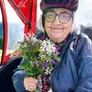 Bildet viser en eldre som sitter i en sykkeltilhenger ute i naturen. I hånden holder hun en bukett markblomster.