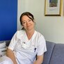 Bildet viser Aina Evensen Helme på jobb i sykepleieruniform. Hun sitter og ser inn i kameraet