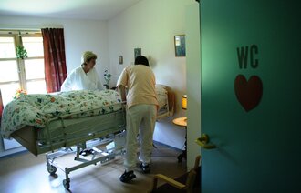 Bildet viser en pasient i en seng på sykehjem, med to pleiere som står ved sengen.