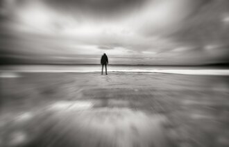 Abstrakt sort-hvitt bilde av en skikkelse som står på en strand og ser ut over havet. 