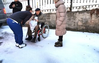 Mann i rullestol, sykepleier lener seg ned og prater. Pårørende til høyre, taxisjåfør til venstre. Vinter i hage. 