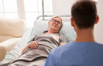 Sykepleier snakker med sengeliggende kvinne.