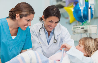 En sykepleier og en lege smiler til en gutt som ligger i en seng på et sykehus.