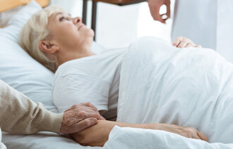 Bildet viser en eldre kvinne som ligger i en seng og en mann som holder henne i armen