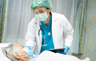 Bildet viser en eldre mann som ligger i senga med maske på, mens legen står ved siden av med maske og visir