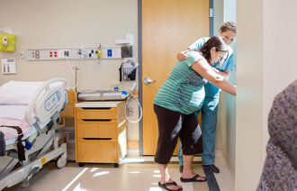Bildet viser en høygravid kvinne som står ved vinduet. En sykepleier holder armen beskyttende rundt henne.