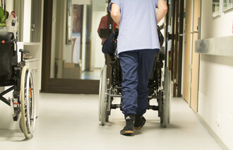 Bildet viser en sykepleier som triller en eldre pasient i rullestol.