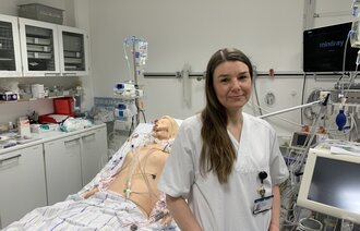 Bildet viser Monica Thallinger foran en sykehusseng, der det ligger en simuleringsdukke.
