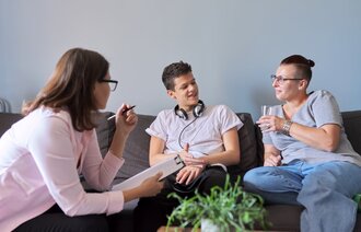 Bildet viser en ung gutt som sitter i en sofa og snakker med to kvinner