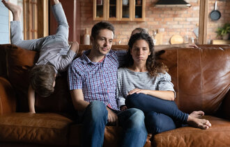 Bildet viser en mann og en kvinne som ser slitne ut og sitter i en sofa, og et barn som klatrer på sofaryggen