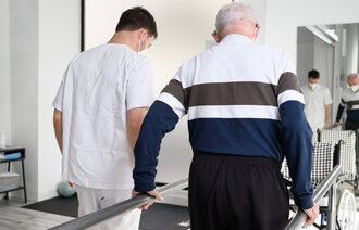 Bildet viser et helsepersonell som hjelper en eldre mann med å øve på å gå 