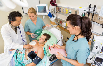 Helsepersonell står rundt sengen til en pasient på sykehus. Sykepleieren skriver i journalen