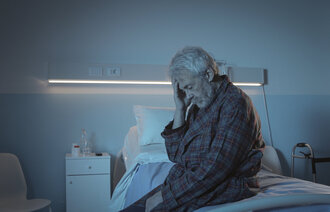 Bildet viser en eldre mann som sitter på sengekanten med hodet i hendene