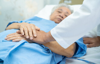 Bildet viser en eldre mann som ligger i en seng, og et helsepersonell som holder vedkommende i hendene