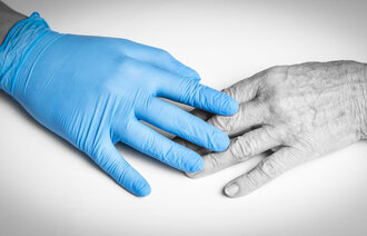 Bildet viser en hånd iført en plasthanske som berører hånden til et eldre menneske