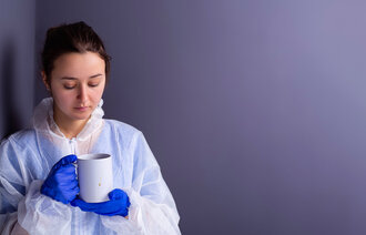 Bildet viser et ungt, kvinnelig helsepersonell i verneutstyr som ser sliten ut og har en kopp i hånden 