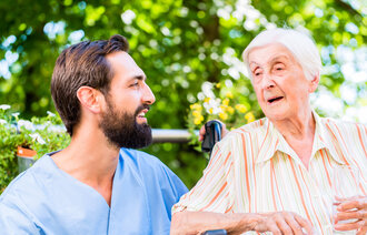 Bildet viser en mannlig sykepleier som snakker med en eldre dame. De sitter ute, og det er god stemning.