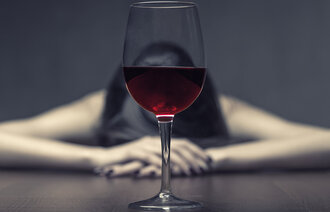 Bildet viser en kvinne som hviler hodet på et bord. Ovenfor henne står et glass rødvin