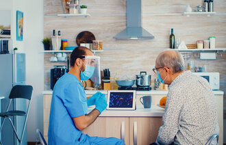 Bildet viser en mannlig sykepleier i blå uniform, munnbind og visir på hjemmebesøk hos en eldre mann, også han med munnbind. De sitter og ser på en ipad.