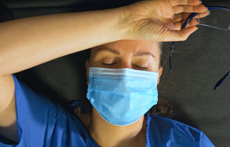 Bildet viser en trøtt sykepleier med munnbind som ligger på gulvet med armen over pannen