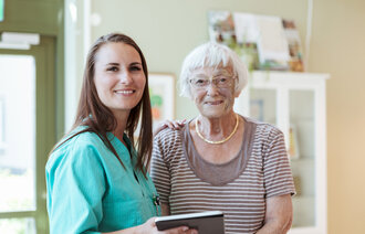 En eldre kvinne og en ung, kvinnelig sykepleier smiler og ser inn i kamera.