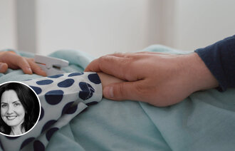 Bildet viser et utsnitt av en voksen hånd som holder rundt en barnehånd. Barnet ligger i sengen.