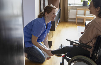 Bildet viser en kvinnelig sykepleier som hjelper en rullestolbruker på med skoene