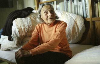 Bildet viser en eldre kvinne som sitter på en seng i hjemmet sitt