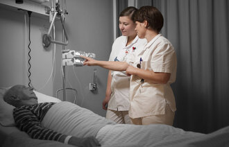 Bildet viser to sykepleiere som står ved en pasientseng