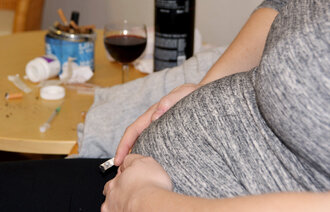 Bildet viser magen til en gravid dame som sitter i en stol. I hånda holder hun en sigarett. På bordet bak står et askebeger fullt av sneiper og et glass med rødvin. Noen pilleglass ligger veltet på bordet.