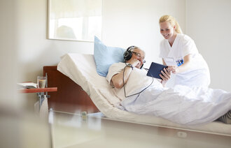 Bildet viser en kvinnelig sykepleierstudent som assisterer en eldre mannlig pasient som ligger i senga, med å bruke et nettbrett
