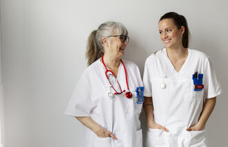 Bildet viser en ung og en eldre sykepleier som smiler til hverandre.