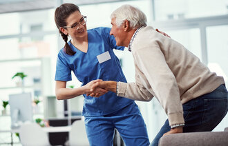 Bildet viser en ung kvinnelig sykepleier som hjelper en eldre mannlig pasient opp fra stolen.