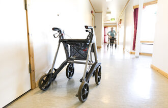Bildet viser korridor med gåstol og en eldre dame som kommer gående - også med gåstol