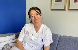 Bildet viser Aina Evensen Helme på jobb i sykepleieruniform. Hun sitter og ser inn i kameraet