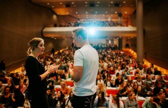 Bildet viser to studenter som står på scenen i et auditorium