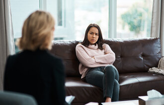 Bildet viser en person som sitter med ryggen til. Overfor henne sitter en ung jente som ser sint ut. Hun har armene i kors og et sint uttrykk