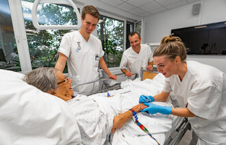 Bildet viser en eldre pasient som ligger i en sykehusseng. Rundt henne står to sykepleierstudenter og en veileder. Alle smiler, og den ene studenten holder pasientens hånd