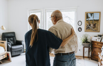 Bildet viser en sykepleier som st&oslash;tter en eldre mann p&aring; sykehjem.