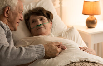 Bildet viser en syk eldre dame som ligger i senga med neseslange. En eldre mann holder henne i hånda og kikker på henne