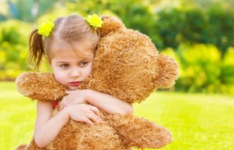 Bildet viser en liten jente som sitter i en eng og klemmer sin kosebamse.