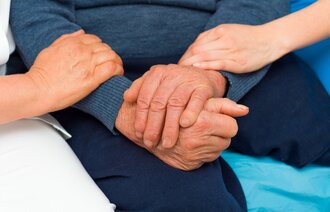 Pasientens to hender er foldet i fanget, mens to hender til pleiere holder varsomt på armene til pasienten. 