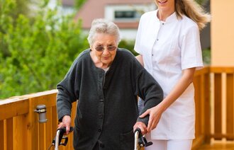 Bildet vise eldre kvinne med gåstol som støttes av av sykepleier.