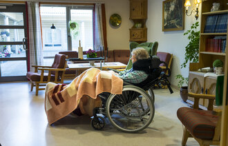 Bildet viser en eldre kvinne liggende i en rullestol i en stue på et behandlingssenter.  