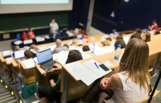 Bildet viser en forelesningssal med mange studenter nedover på radene. Foran står foreleseren.
