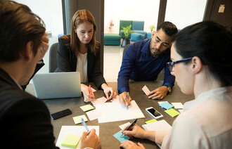 Bildet viser gruppearbeid på et kontor, der flere mennesker sitter rundt et bord med PC, ark og gule lapper foran seg.