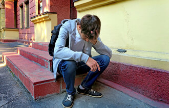 Bildet viser en tenåringsgutt med ransel som sitter på en trapp utenfor skolen. Han er lei seg.